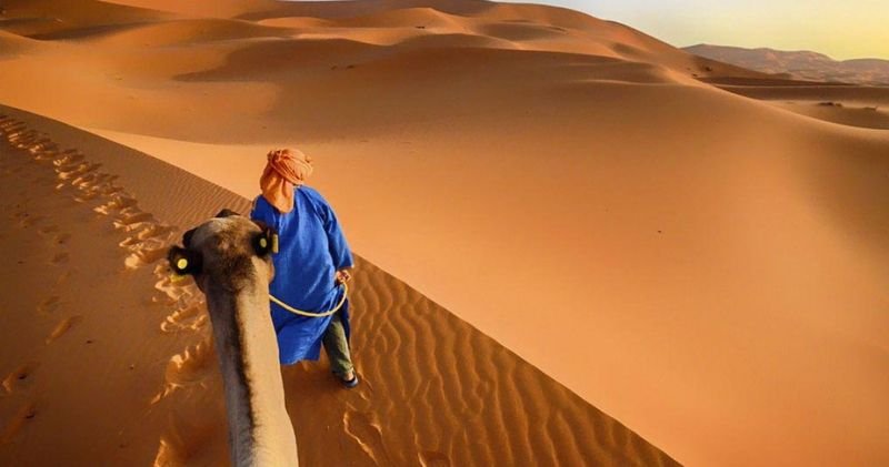 Homme en robe bleue menant un dromadaire sur une dune