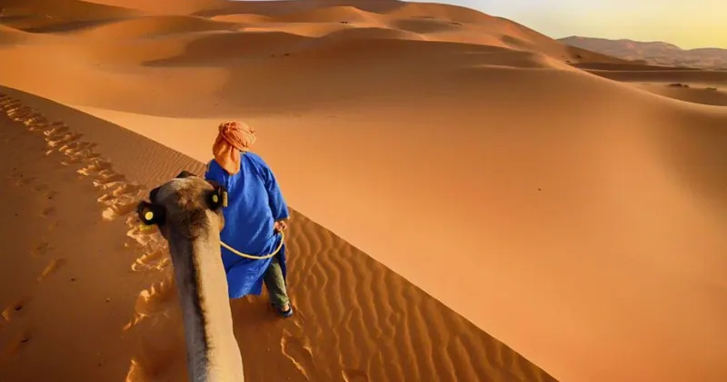 Homme en robe bleue menant un dromadaire sur une dune