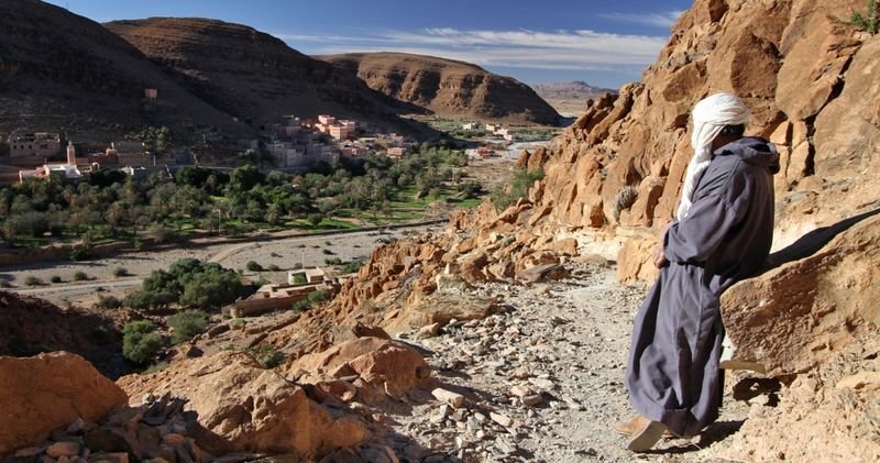 Homme en djellaba sur un versant rocheux dominant un village de vallée